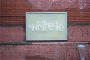Red brick wall with a blank plastic frame that awaits an advertising poster, but has the words "white lie" sprayed on it in white paint